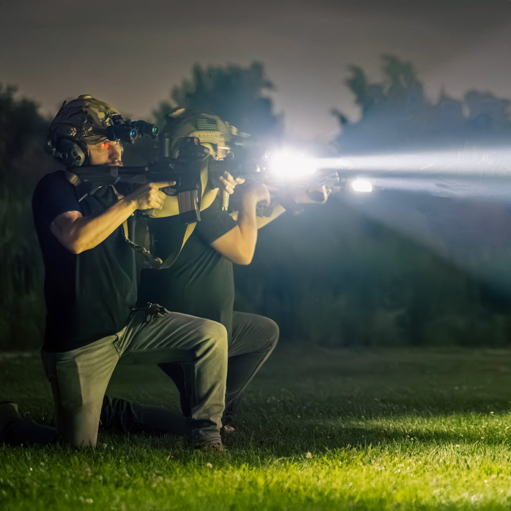 Two man kneeling in a field pointing rifles with flashlights attached.
