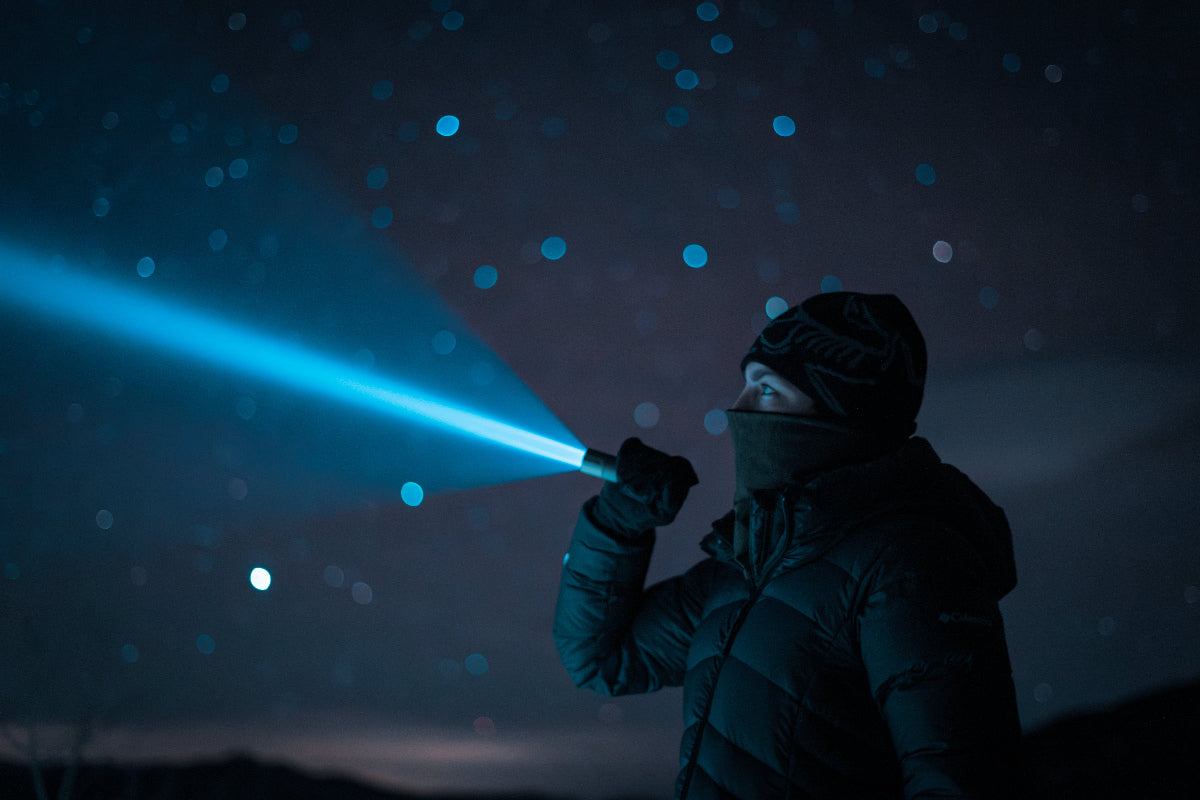 woman in a coat and beanie shinning flashlight with night sky in background.