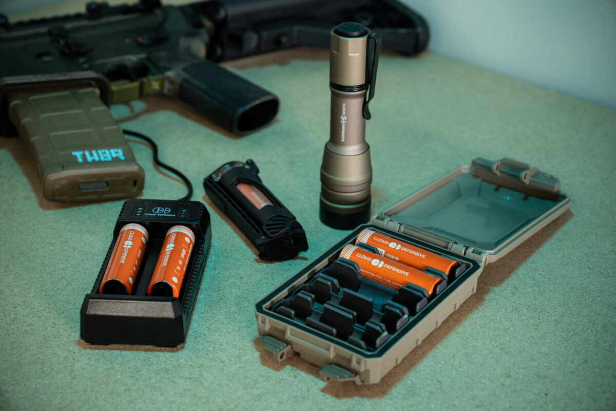 FDE MCH Duty flashlight on table surrounded by extra batteries in a case, a battery charger and rifle in the background.